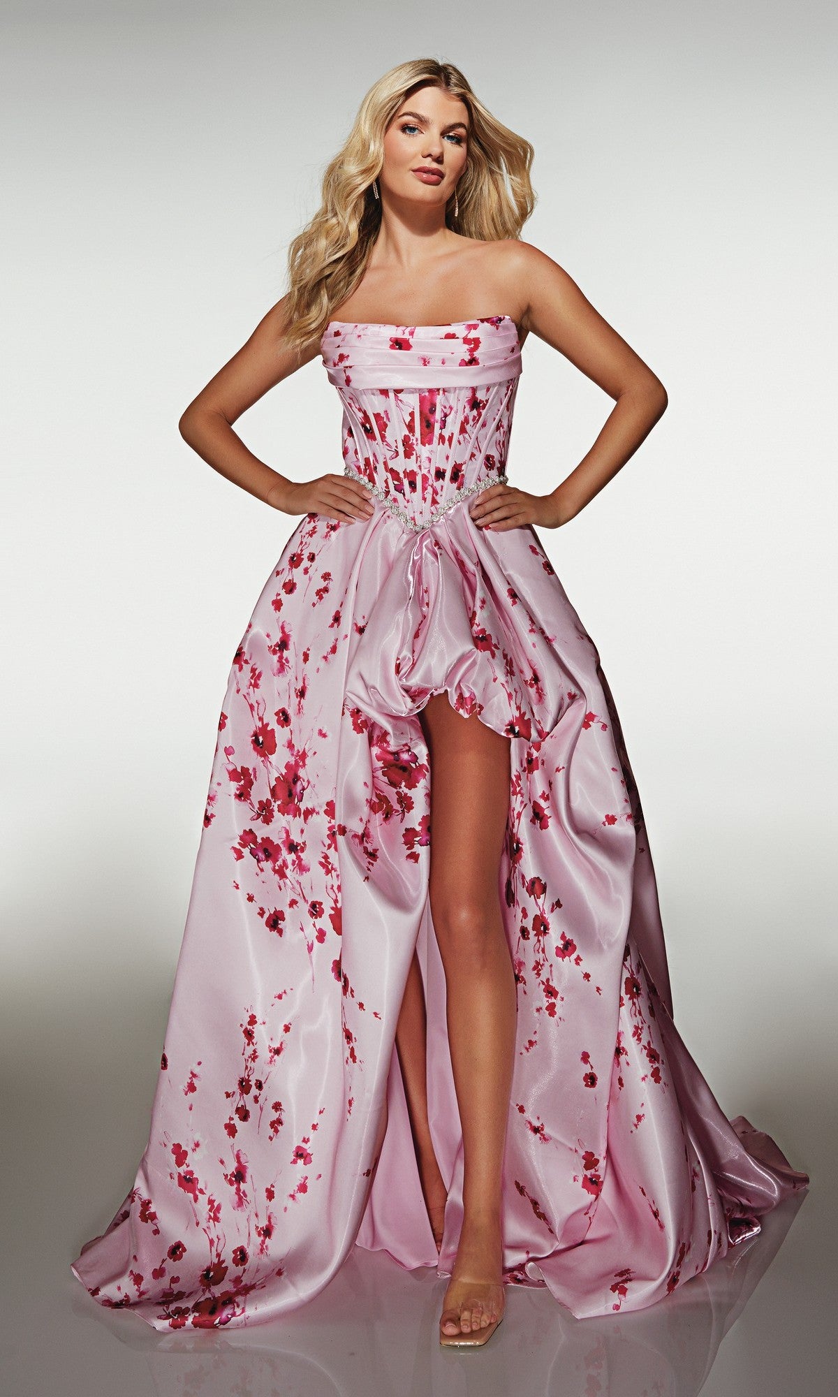 A woman stands with hands on hips, wearing the Alyce 61989 long prom dress—a strapless satin ball gown in light pink with red floral patterns and a high-leg slit.